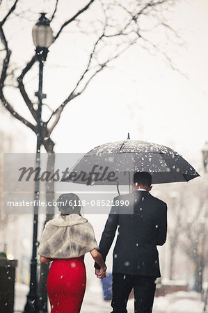 A woman in a long red evening dress and a man in a suit, walking through snow in the city.