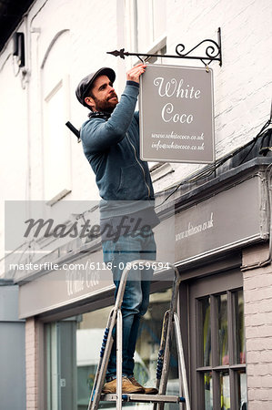 A man on a ladder fixing a painted name sign onto a bracket on a shopfront.