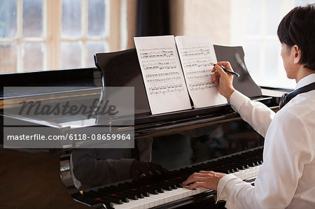 Young man sitting at a grand piano in a rehearsal studio, annotating sheet music.