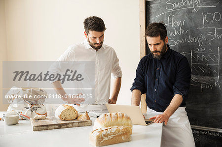 Two bakers standing at a table, using a laptop computer, freshly baked bread, a blackboard on the wall.