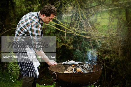 Chef standing in a garden, grilling a fish on a barbecue.