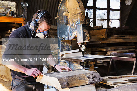 Man working in a carpentry workshop, wearing a respirator and hearing protector, measuring a plank of wood.