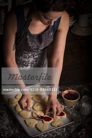Valentine's Day baking, young woman standing in a kitchen, spreading raspberry jam on heart shaped biscuits.