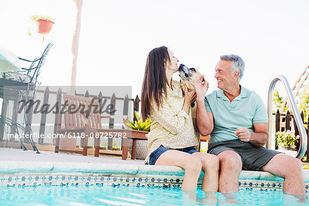 A couple sitting on the edge of a swimming pool, with their dog.