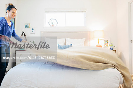 A woman spreading a plain quilt across a double bed  in a light airy apartment bedroom.