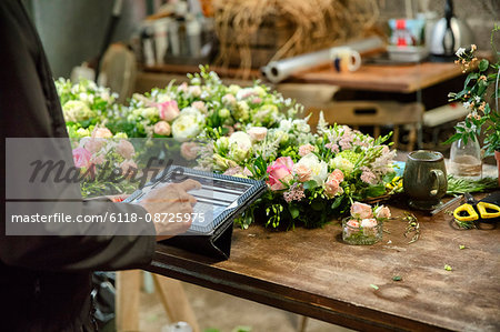 A woman in a florist's workshop using a digital tablet, at a workbench with flower arrangements.