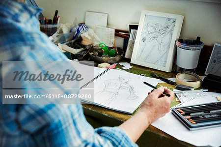 An artist working, holding a pencil over a sketch in progress in an open sketchbook.