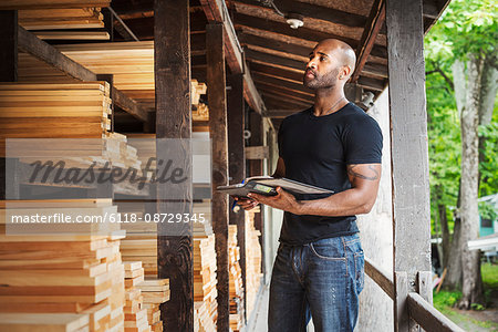 Man standing in a lumber yard, holding a folder, checking notes.