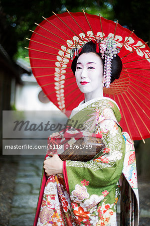 A woman dressed in the traditional geisha style, wearing a kimono and obi, with an elaborate hairstyle and floral hair clips, with white face makeup with bright red lips and dark eyes holding a red paper parasol.