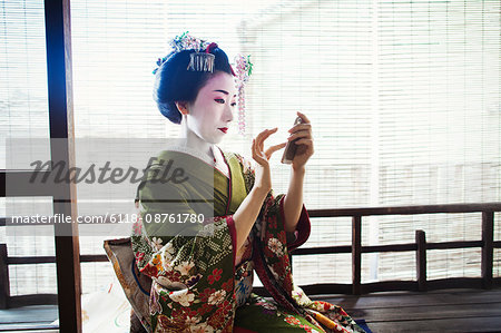 A woman dressed in the traditional geisha style, wearing a kimono and obi, with an elaborate hairstyle and floral hair clips, with white face makeup with bright red lips and dark eyes taking a selfie of herself.