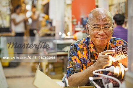 A senior craftsman in a glass maker's workshop sitting at a workbench using a light to check an etched or cut glass pattern.