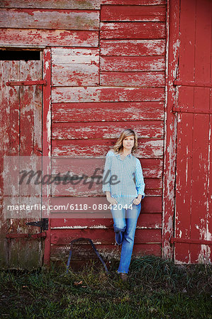 A young woman in jeans leaning against a red painted barn building.