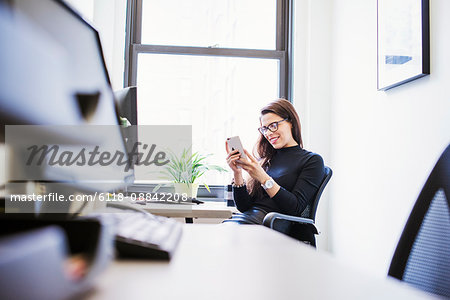 A young woman sitting at a desk in an office looking down at a cellphone.