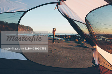 Man framed by camping tent, standing on beach and looking through binoculars at dusk, Olympic National Park, Washington, USA.