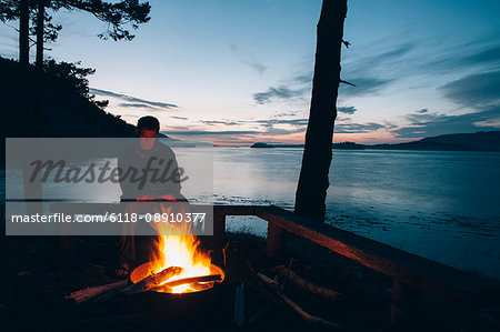 Man sitting by campfire at dusk, San Juan Islands in the distance, Washington, USA.
