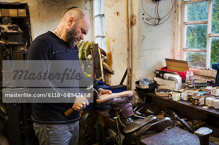 Man standing in a shoemaker's workshop by a window, using a hammer on the soles of a work boot.