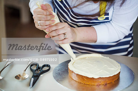 Close up of person wearing a blue and white stripy apron, holding a piping bag, decorating a cake with cream, spoons and scissors on table.