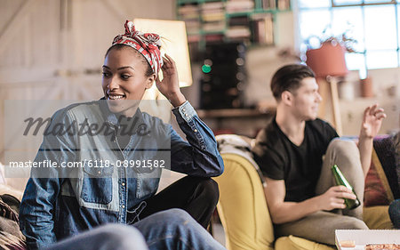 Young woman wearing headscarf and young man holding beer bottle sitting indoors on a sofa, smiling.