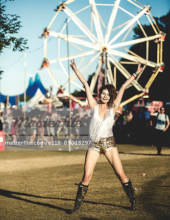 Young woman at a summer music festival wearing golden sequinned hot pants, face painted, smiling at camera, arms raised.