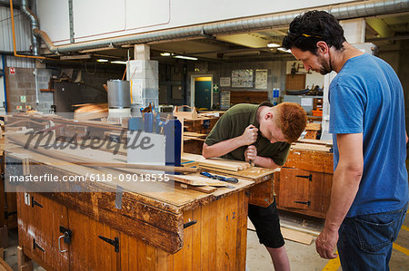 Two men standing at a workbench in a boat-builder's workshop, working on wooden joint.