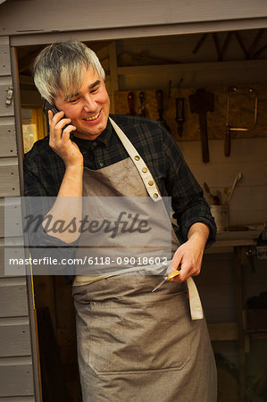 Man with grey hair wearing a carpenter's apron standing in doorway of shed, talking on a mobile phone