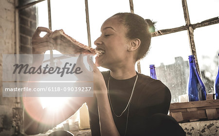Smiling young woman sitting indoors by a window, eating slice of pizza.