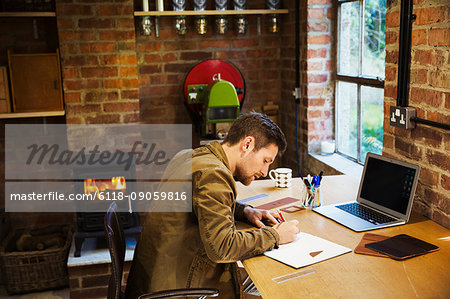 A designer seated in his leatherwork workshop, at a desk drawing on paper. A woodburning stove with a lit fire.