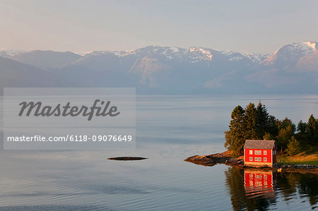 Traditional red wooden house standing on sea shore, snow capped mountains in the distance.