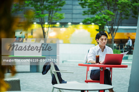 Businessman wearing white shirt sitting outdoors at red table, working on laptop.