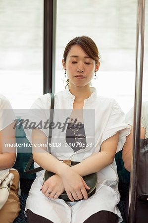 Woman with black hair wearing white shirt sitting on commuter train, eyes closed.