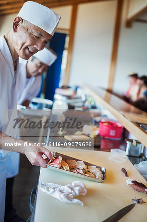 Two chefs working at a counter at a Japanese sushi restaurant, holding metal tray with fresh fish and seafood.