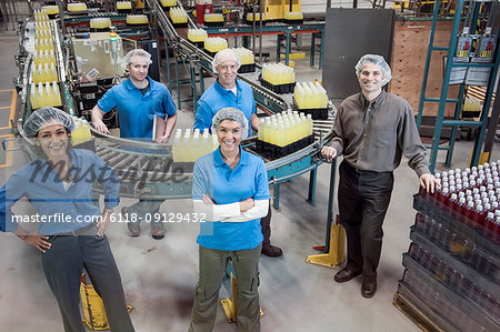Portrait of a team of male and female workers and male management person standing next to a production line conveyor belt of lemon flavoured water  in a bottling plant.