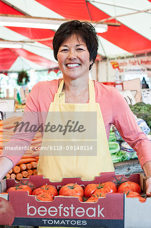 Smiling woman wearing apron holding large tray of fresh tomatoes at a fruit and vegetable market.