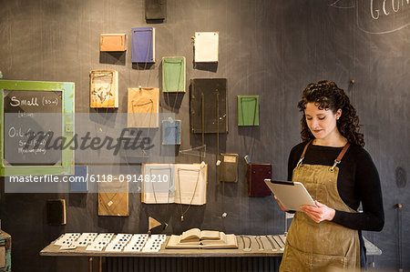 Woman with curly brown hair wearing apron standing in her pottery shop, looking at digital tablet, display of books and jewellery.