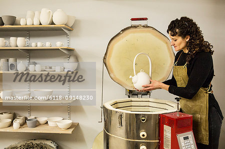 Woman with curly brown hair wearing apron standing in pottery workshop, placing vase into kiln.