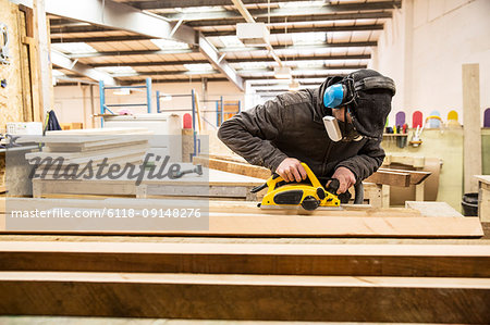 Man wearing ear protectors, protective goggles and dust mask standing in a warehouse, sanding planks of recycled wood.