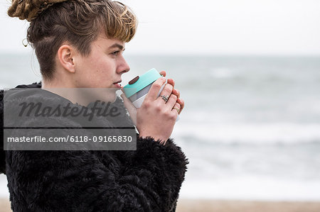 Portrait of young woman with brown hair and dreadlocks and a lip piercing wearing black furry jacket, standing by ocean, holding mug to her lips.