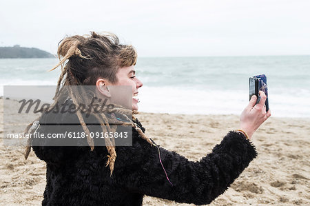 Smiling young woman with brown hair and dreadlocks and a lip piercing wearing black furry jacket, sitting on sandy beach by ocean, taking selfie with mobile phone.