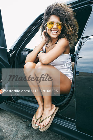Portrait of smiling young woman with brown curly hair sitting in car, wearing sunglasses, white vest and flip flops.