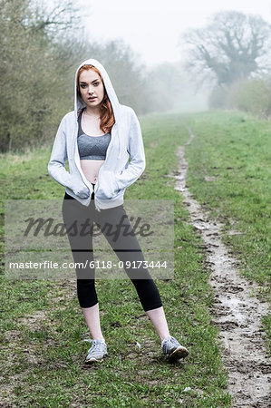 Young woman with long red hair wearing sports kit, exercising outdoors, looking at camera.