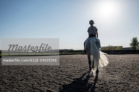 Rear view of teenage girl riding a grey horse in a paddock.