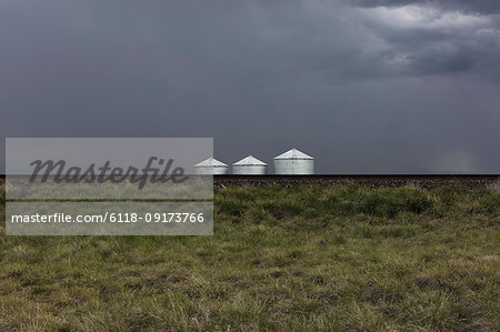 Grain silos and storm clouds over vast farmland and prairie, train tracks in foreground