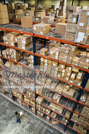A view from above looking down on a warehouse worker checking inventory of boxes on racks in a distribution warehouse.