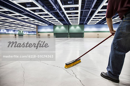 A low view closeup of a man sweeping the floor of a convention centre arena.