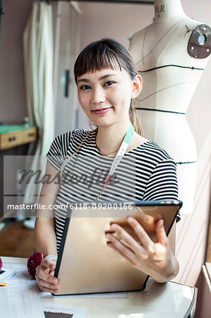 Japanese female fashion designer working in a studio, holding digital tablet, smiling at camera.