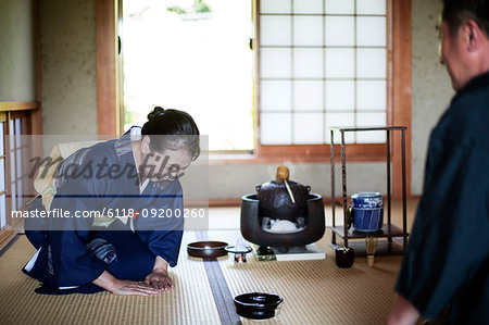 Japanese woman wearing traditional bright blue kimono with cream coloured obi and man kneeling on floor during tea ceremony.