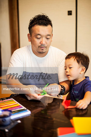 Japanese man and little boy sitting at a table, making Origami animals using brightly coloured paper.