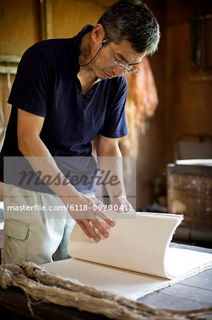 Japanese man standing in a workshop,  checking through sheets of traditional Washi paper.