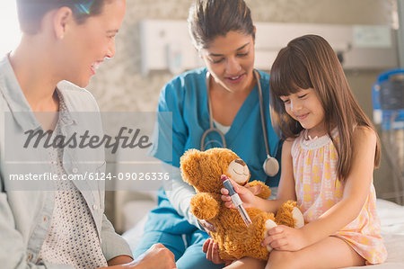 Female nurse and mother watching girl patient using insulin pen on teddy bear