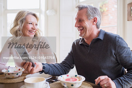 Laughing mature couple using smart phone and eating breakfast